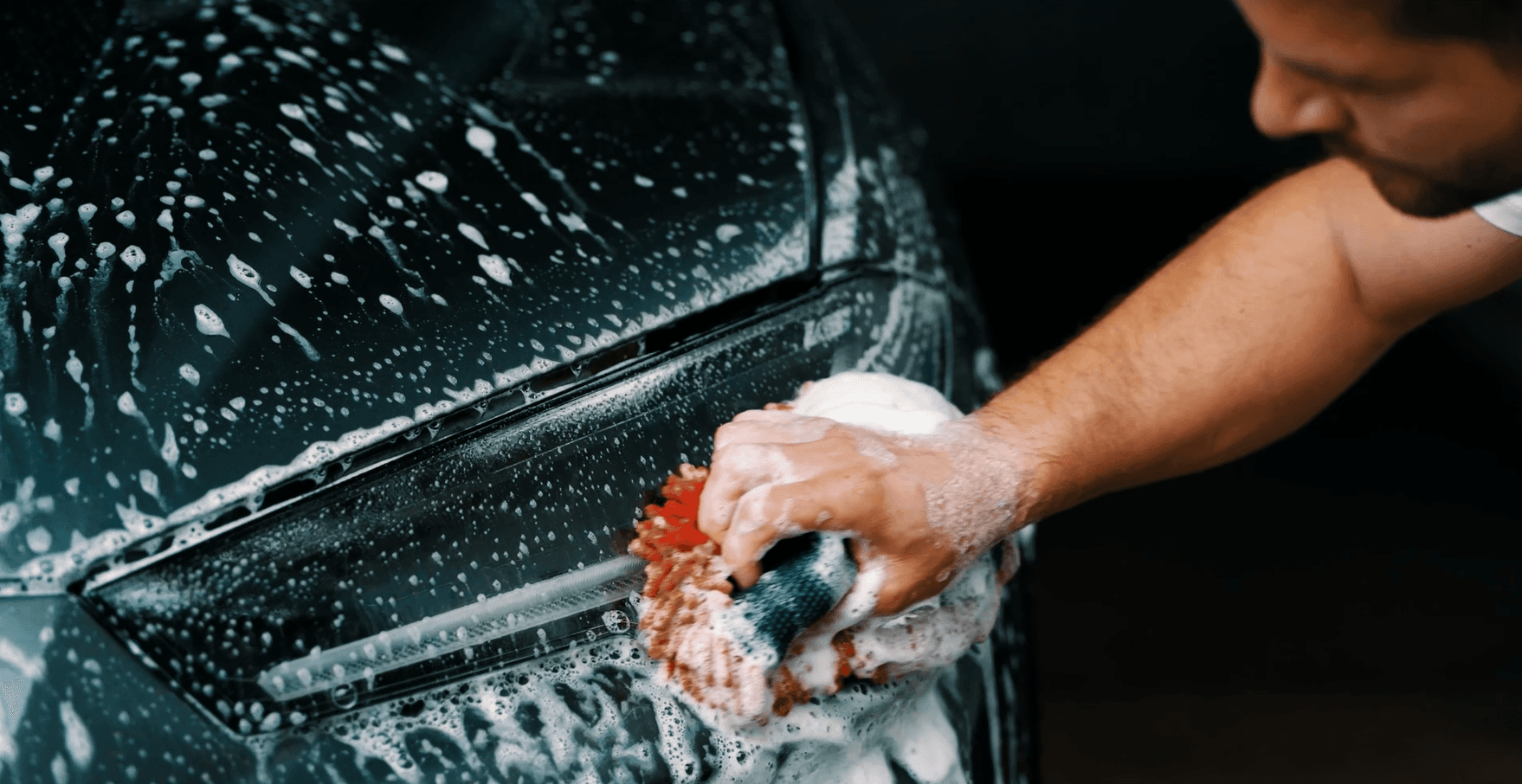 A man washing the front of a gray sports car with a wash mitt.